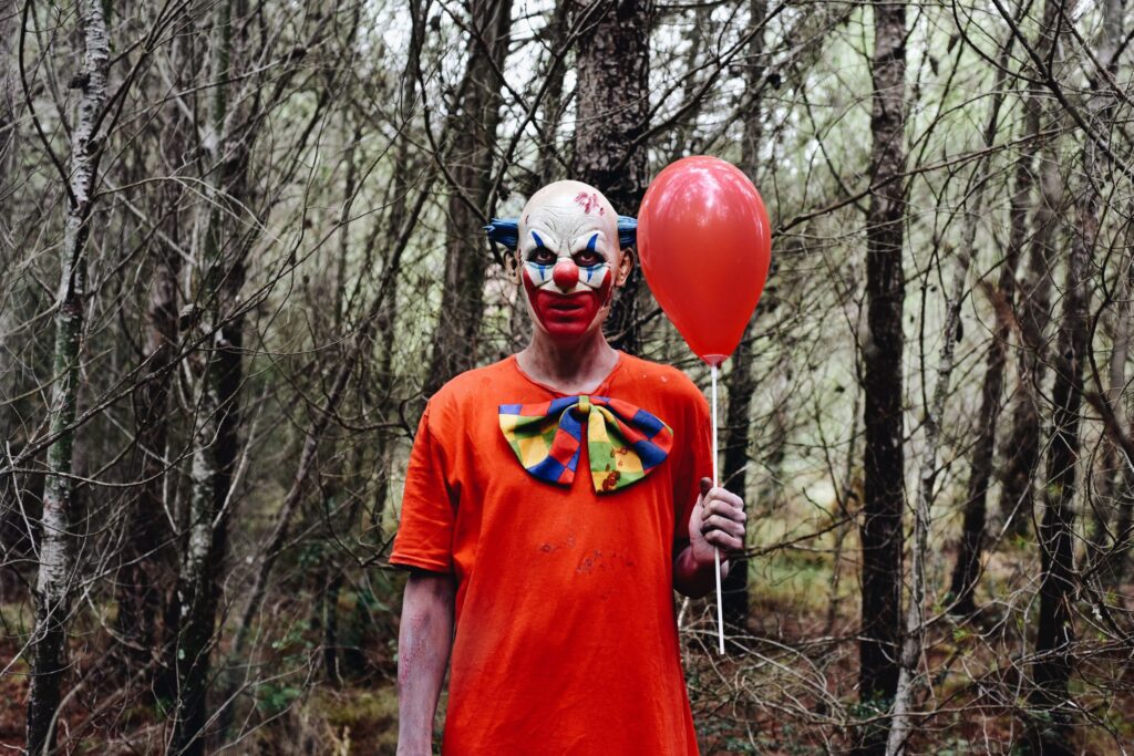 a scary evil clown wearing a dirty costume, holding a red balloon in his hand, in the woods